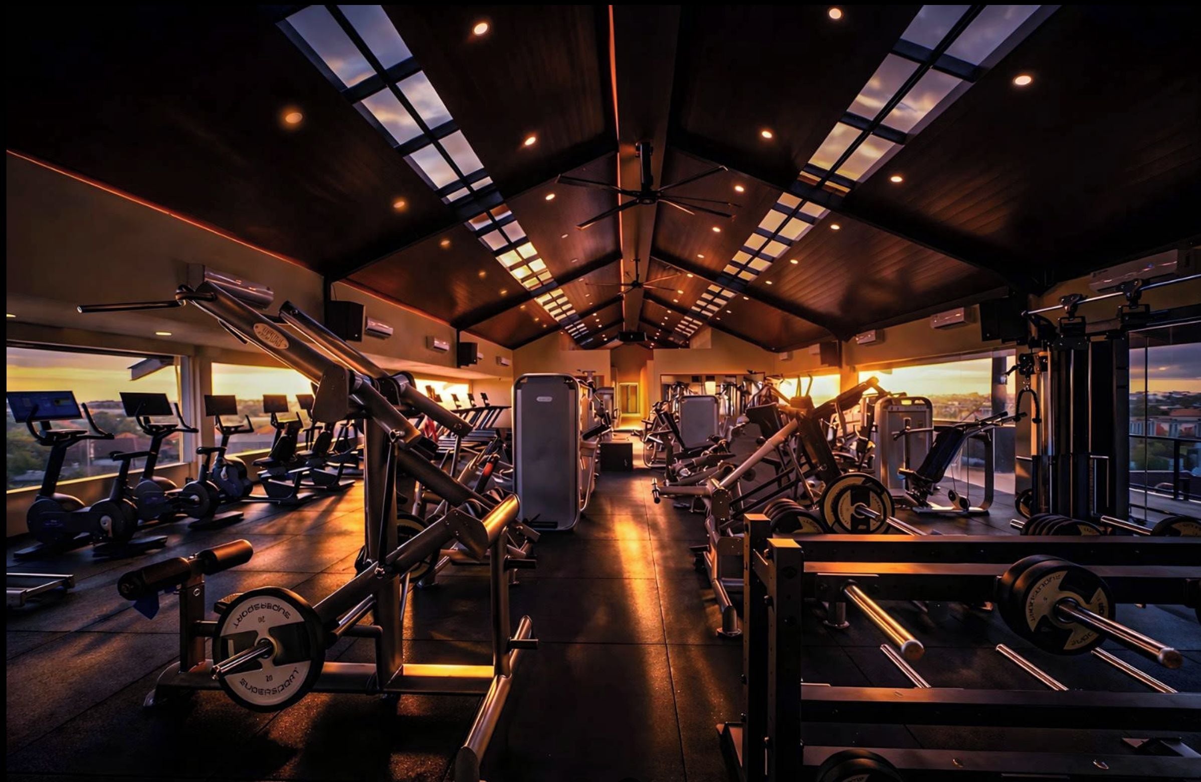 Un gimnasio con diversos equipos de ejercicio, ventanas amplias y un techo alto.