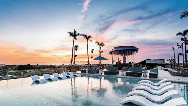 Ein Pool mit Infinity-Kante auf dem Dach, mit Liegestühlen und Sonnenschirmen, Palmen und Blick auf den Sonnenuntergang.
