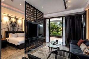 Quarto de hotel moderno com uma cama grande, televisão de tela plana, sofá, mesa pequena e varanda com cortinas.