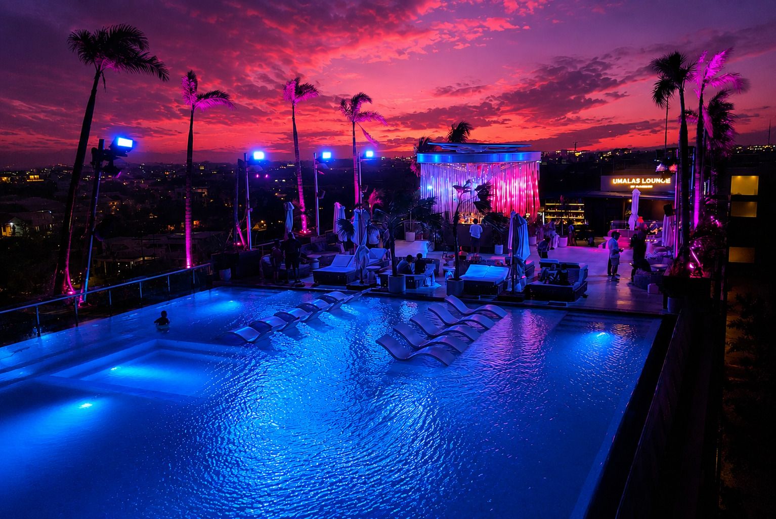 A rooftop pool area with lounge chairs and umbrellas, a pool with blue lighting, and a view of the city skyline at dusk.