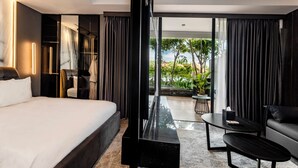 Ein modernes Hotelzimmer mit einem großen Bett, einer Sitzecke mit Sofa und Couchtisch, einem kleinen Tisch mit Lampe und Blick ins Freie.