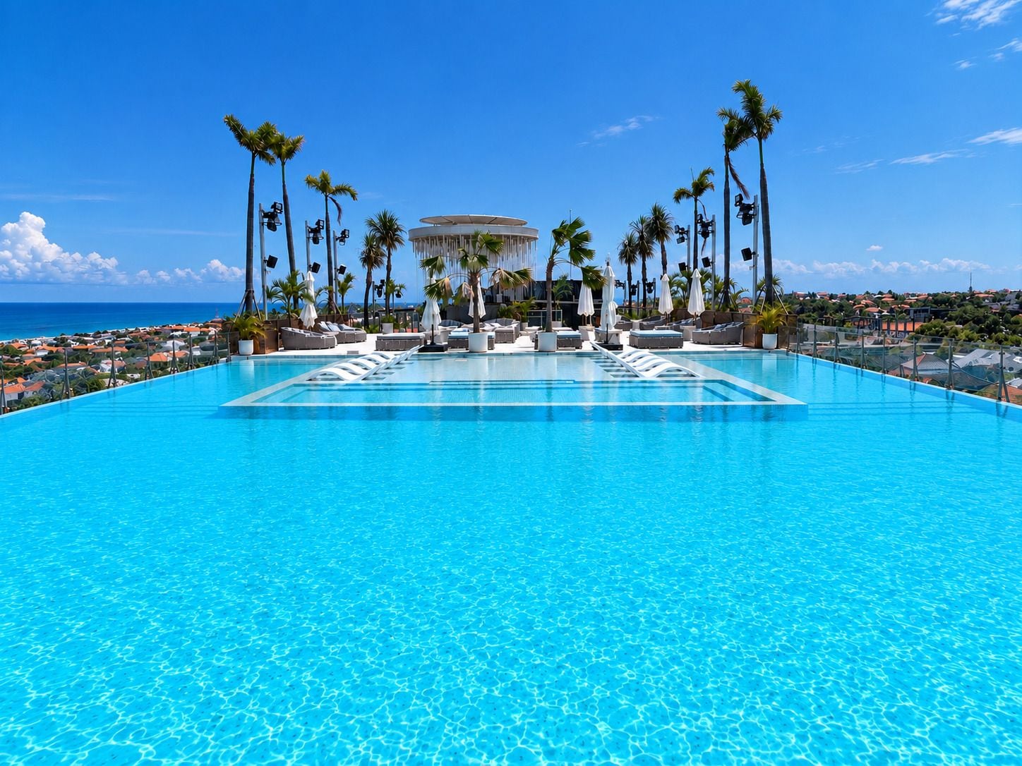 A large, clear blue pool with lounge chairs and umbrellas, surrounded by palm trees and a view of the ocean.