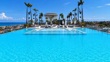 A large, clear blue pool with lounge chairs and umbrellas, surrounded by palm trees and a view of the ocean.
