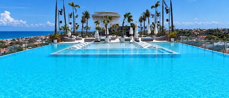 A large, clear blue pool with lounge chairs and umbrellas, surrounded by palm trees and a view of the ocean.