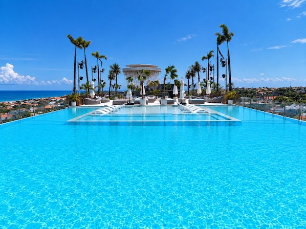 Una grande piscina cristallina con lettini e ombrelloni, circondata da palme e vista sull'oceano.