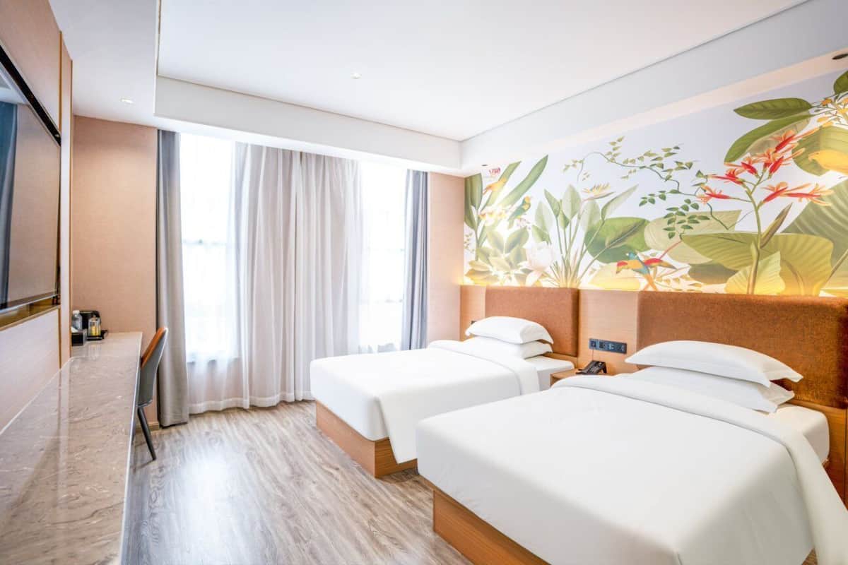 A hotel room with two beds, a large window with curtains, a dining area with a table and chairs, and a mural with tropical plants.