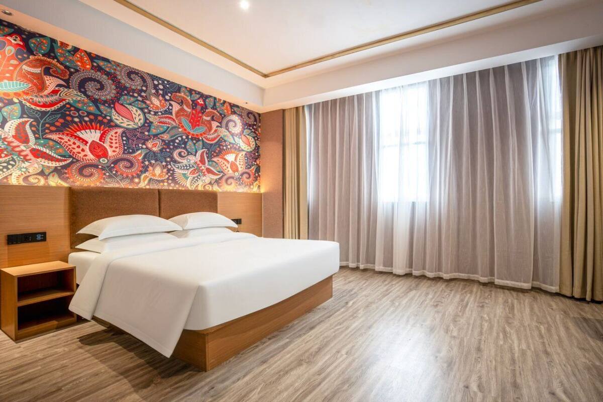 A modern hotel room with a large bed, a nightstand, a wall-mounted clock, and a large window with curtains.
