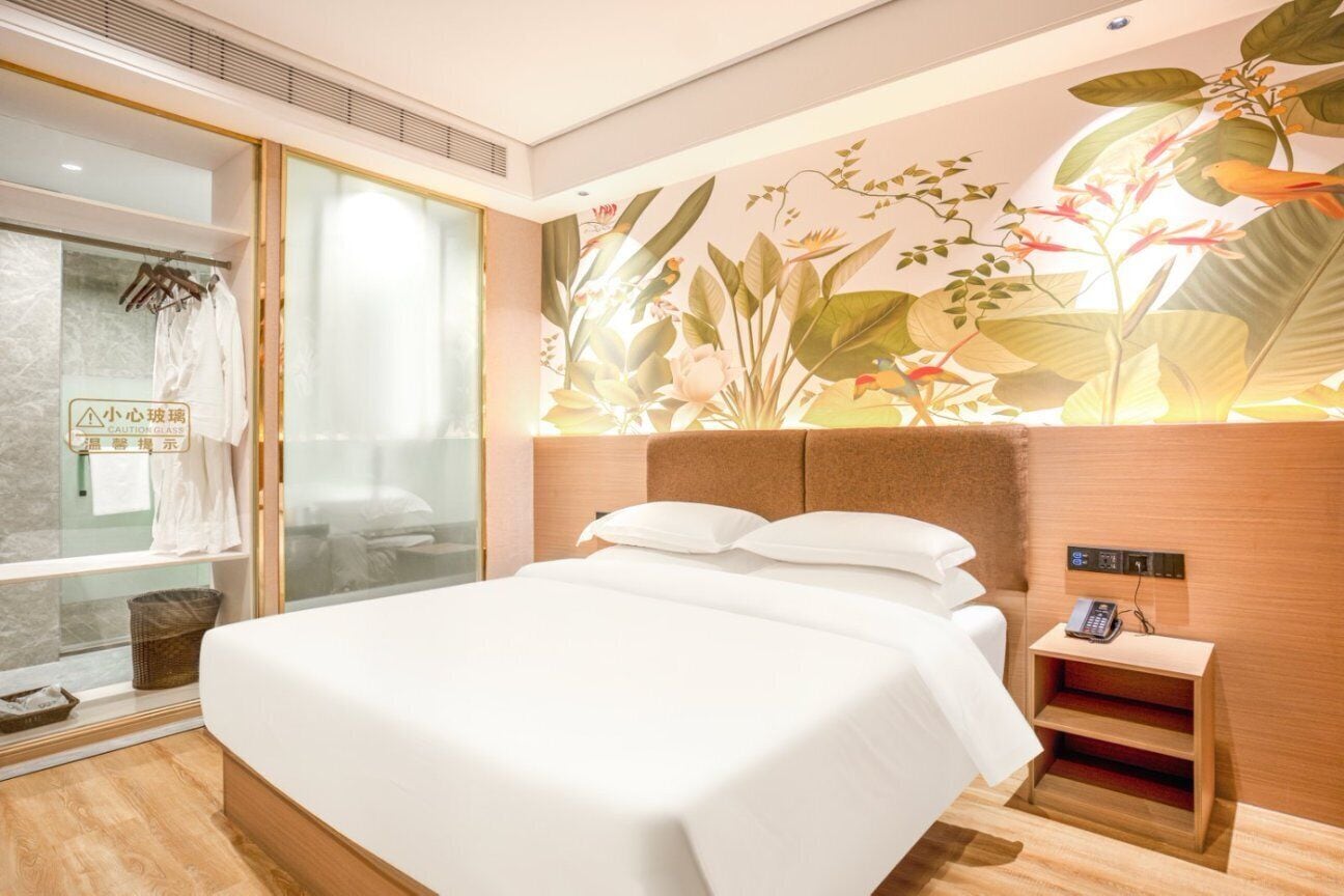 A hotel room with a large bed, a bedside phone, a glass shower enclosure, and a wall with a tropical plant mural.