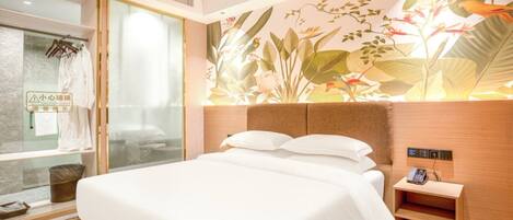 A hotel room with a large bed, a bedside phone, a glass shower enclosure, and a wall with a tropical plant mural.