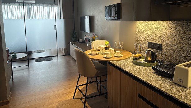 A modern kitchen with a dining area, featuring a granite countertop, a built-in microwave, and a flat-screen TV. - The Vilton (Raipur)
