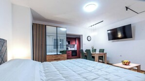 Ein modernes Hotelzimmer mit einem großen Bett, einer kleinen Essecke, einem an der Wand montierten Fernseher und einem Fenster mit Jalousien.