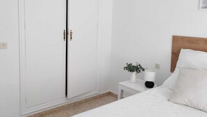 Une chambre à coucher avec un lit, une table de chevet ornée d’une plante, un luminaire suspendu en osier et une armoire blanche.