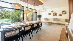 Un comedor moderno con una mesa larga, sillas negras y lámparas colgantes tejidas. Se ve un sofá y una pequeña mesa al fondo.