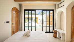 Une chambre dotée d’un grand lit, d’une porte en bois, d’une porte-fenêtre coulissante donnant sur une piscine et d’un banc intégré avec une table d’appoint.