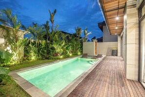 Egy úszómedence, melynek köré fából készült napozóteraszot építettek, és amelyet buja zöld növényzet, valamint egy modern épület vesz körül.
