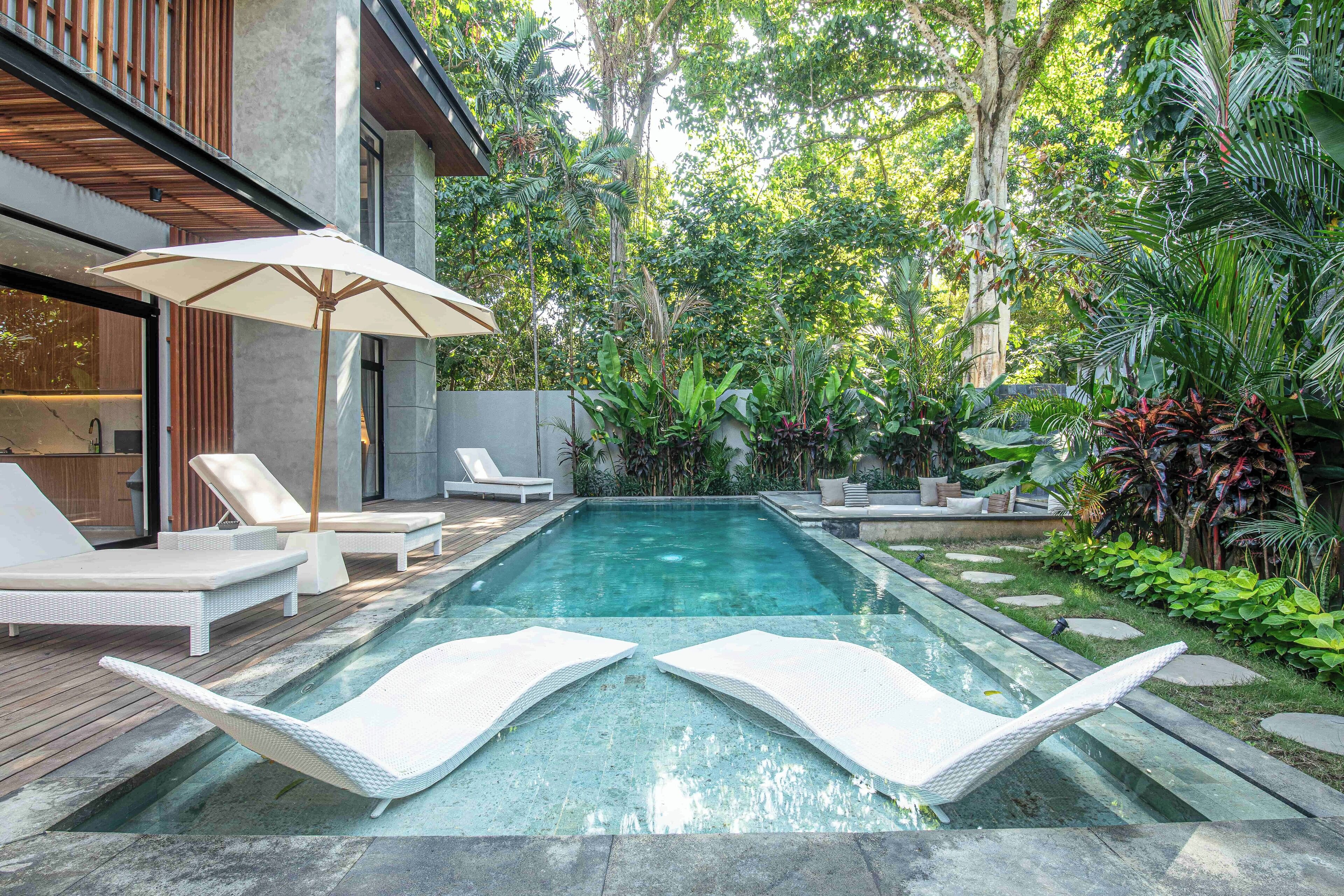 En pool med liggestole og parasol, omgivet af frodig beplantning og en moderne bygning.