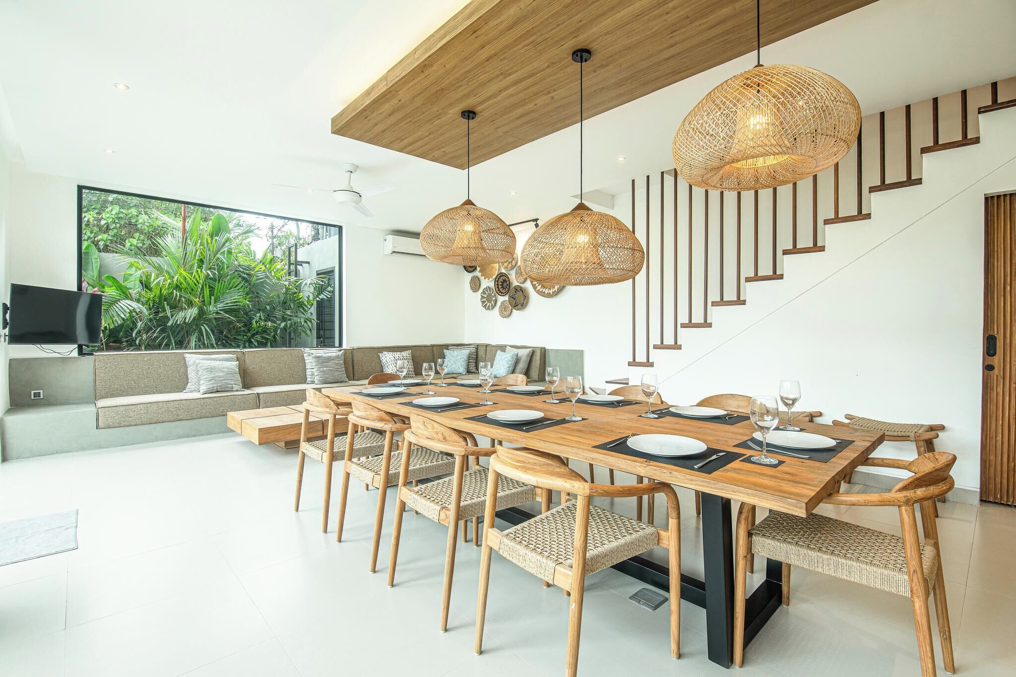 Un comedor moderno con una mesa de madera dispuesta para seis personas, rodeada de sillas de mimbre. En el fondo, se observa una escalera con escalones de madera y una pared blanca.