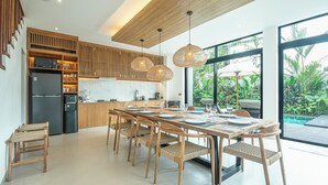 A modern kitchen with wooden cabinetry, a dining area with a long table and chairs, and large windows offering a view of the outdoors. - Elegant 5BR Modern Villa Steps From Caf Del Mar (Seminyak)