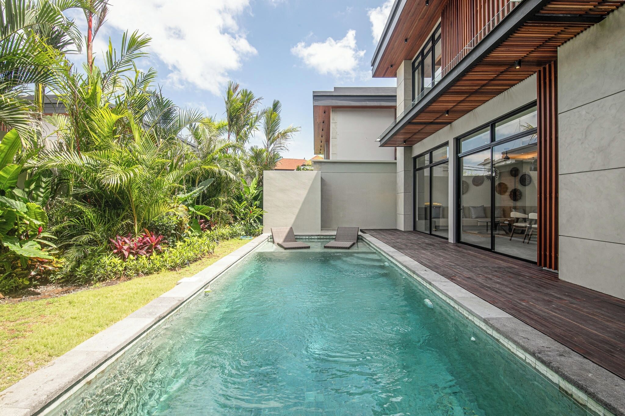 A modern outdoor pool area with a clear pool, wooden deck, and lush tropical plants.