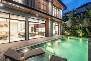 A modern house with a swimming pool, wooden deck, and glass doors leading to a dining area. - Exquisite 5BR Canggu Villa Steps From the Beach (Canggu)