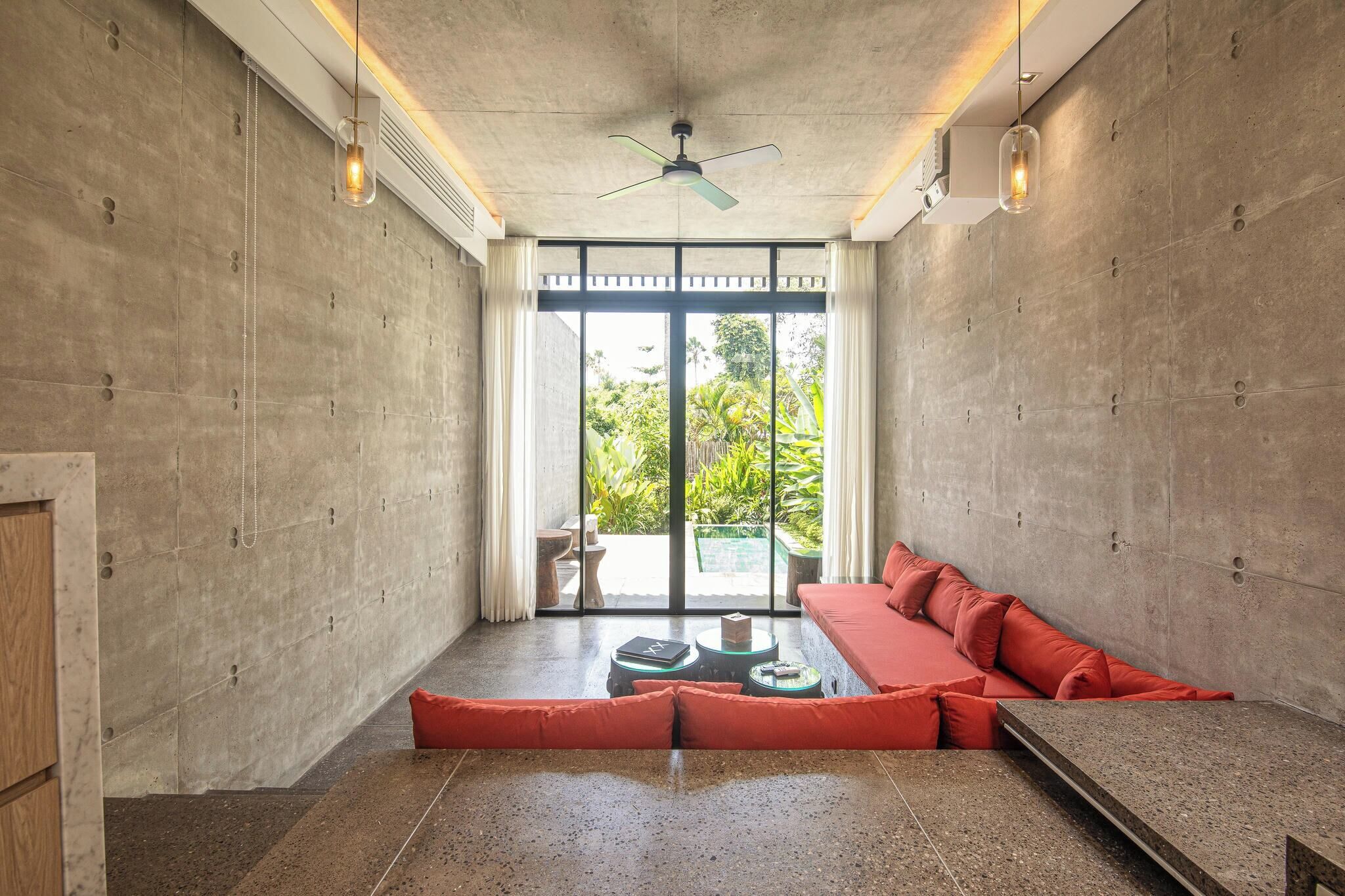 Un intérieur moderne avec un mur en béton, un canapé rouge, un ventilateur de plafond et une vue sur un espace extérieur, accessible par de grandes portes vitrées.
