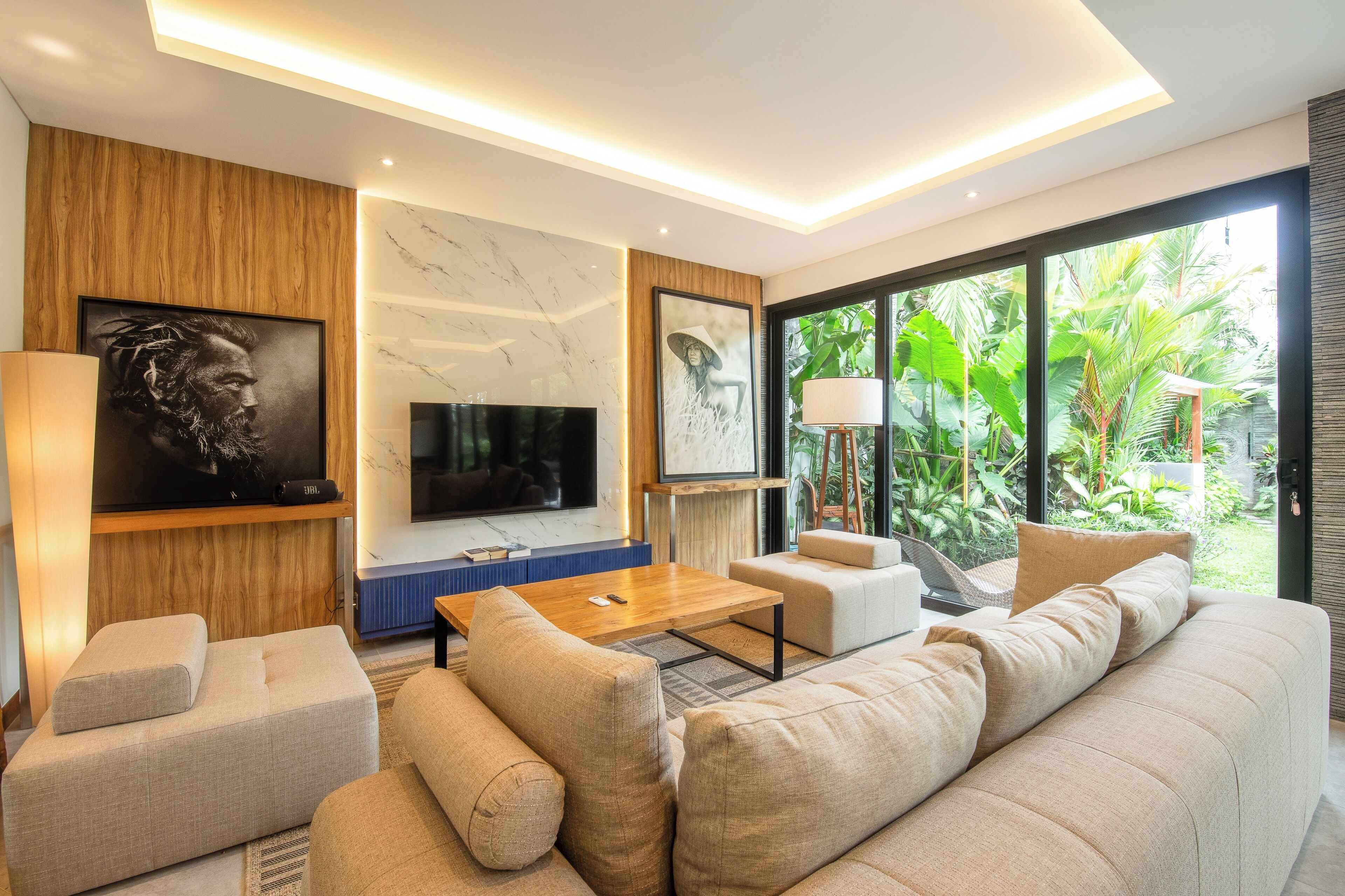 Een moderne woonkamer met een grote bank, een salontafel en een flatscreen televisie. Er hangen kunstwerken in de lijsten aan de muren en er is uitzicht op groen buiten.