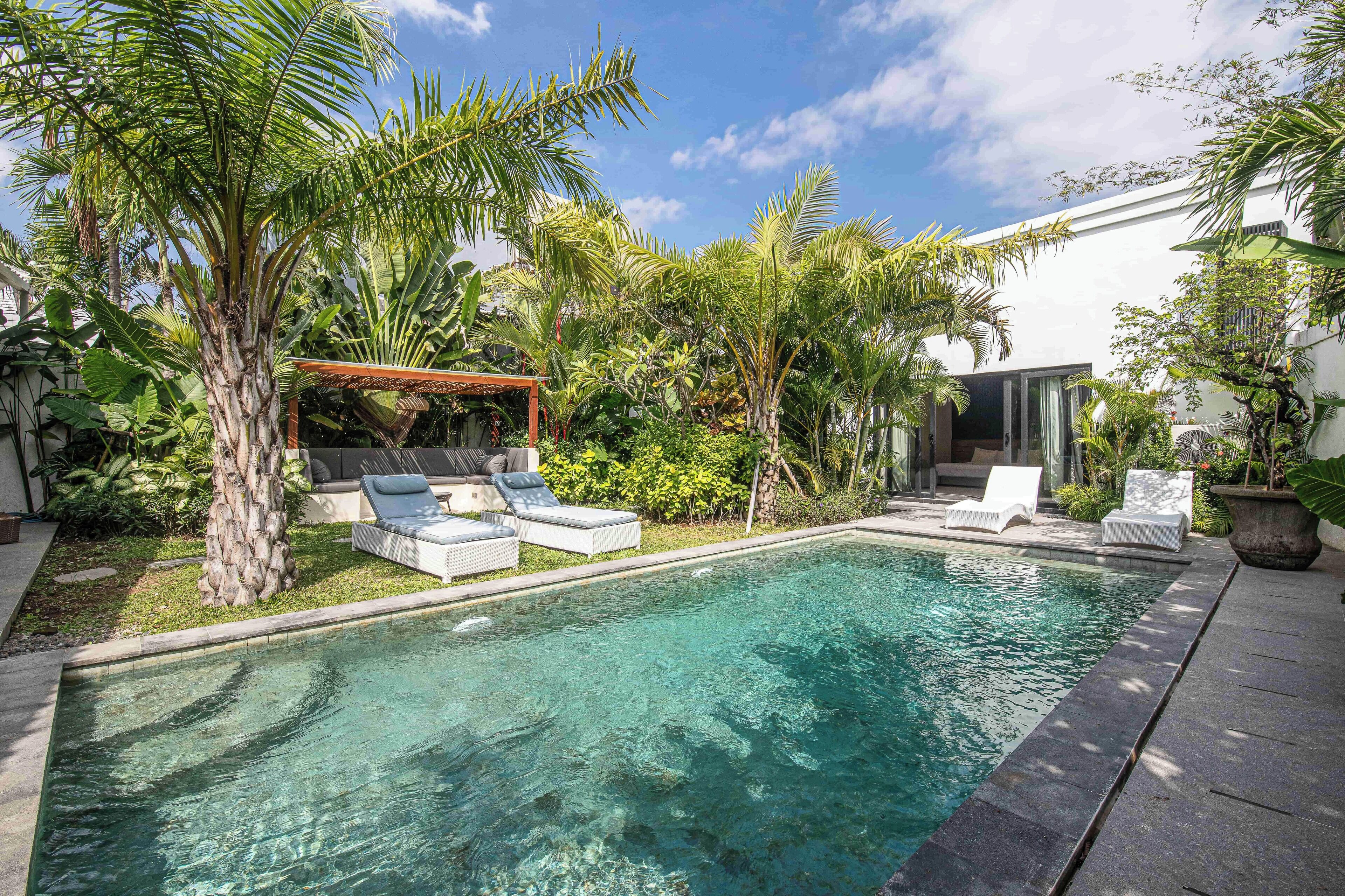 Una piscina con sillones de descanso, rodeada de plantas tropicales y una pérgola.