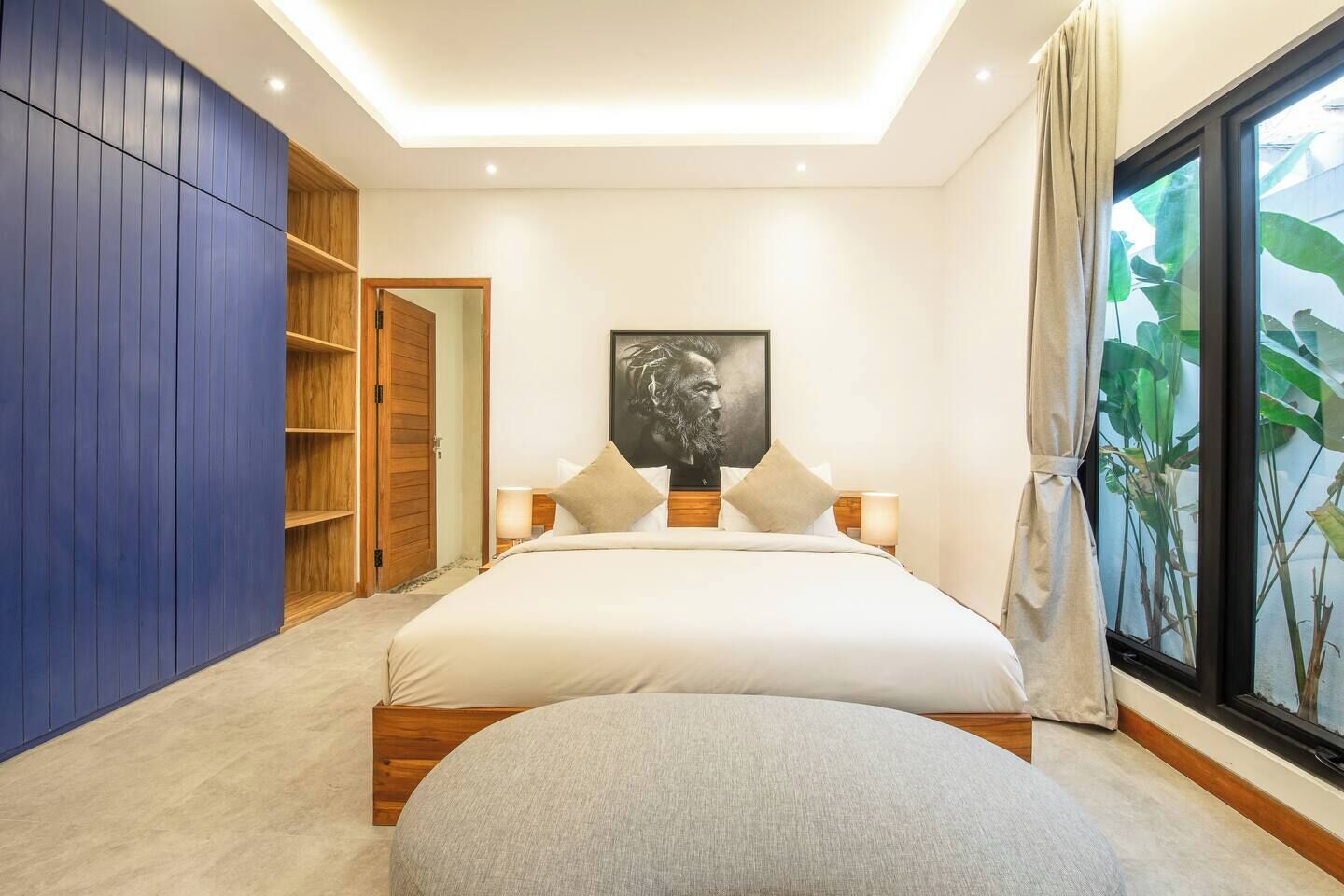 Ein Schlafzimmer mit einem großen Bett, einem Holzschrank, einem Nachttisch mit Lampe und einem großen Fenster mit Blick auf Pflanzen.