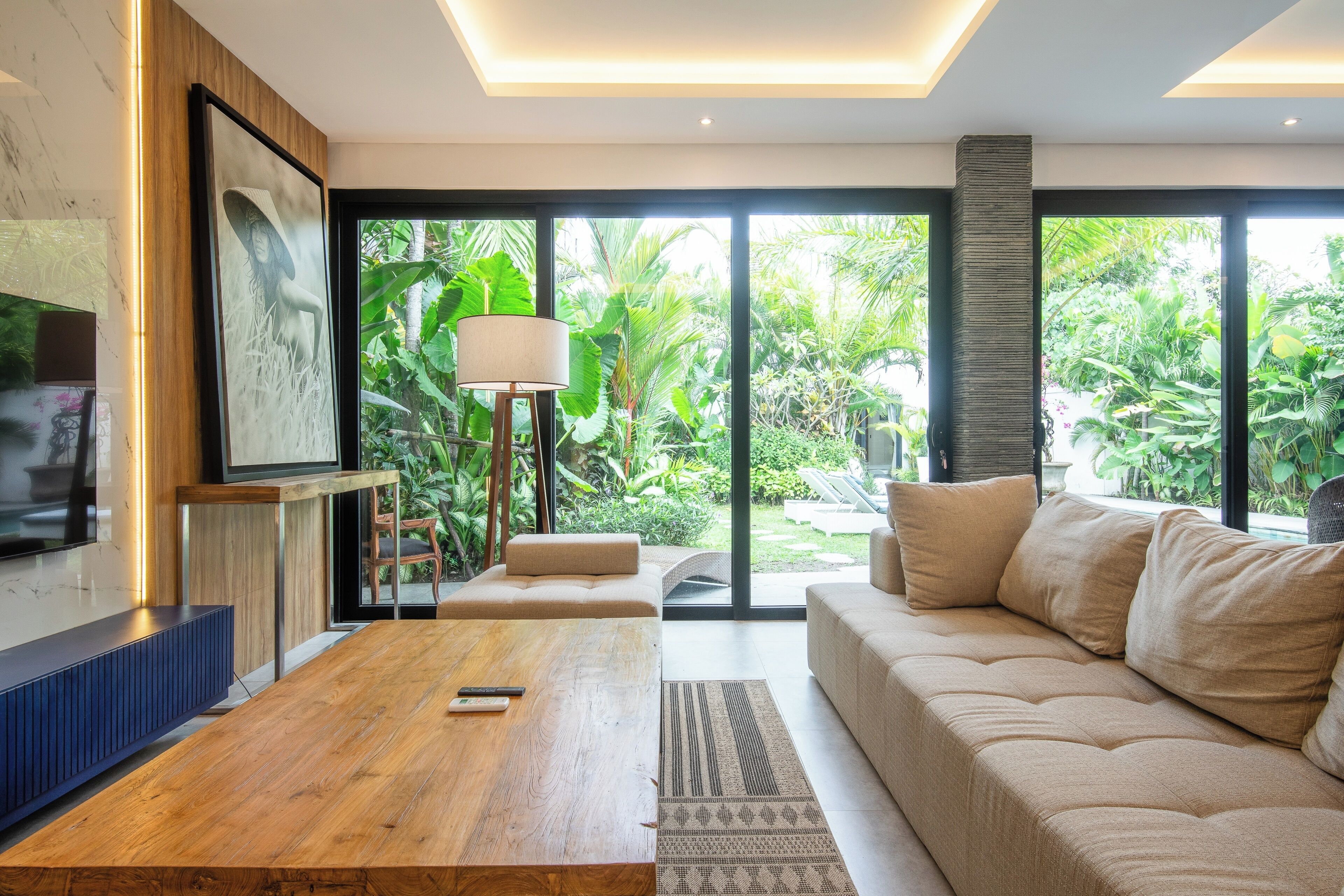 Een moderne woonkamer met een houten salontafel, een beige bank en een groot raam met uitzicht op weelderige begroeiing.