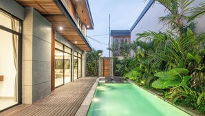 A modern outdoor pool area with a wooden deck, surrounded by lush tropical plants. - Sophisticated 5-br Canggu Retreat Near Beach (Canggu)
