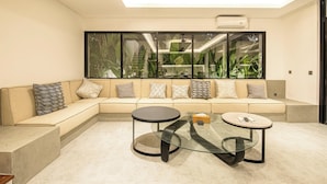 A modern living room with a beige sectional sofa, two glass-top coffee tables, and a large window with a view of greenery. - Sophisticated 5-br Canggu Retreat Near Beach (Canggu)