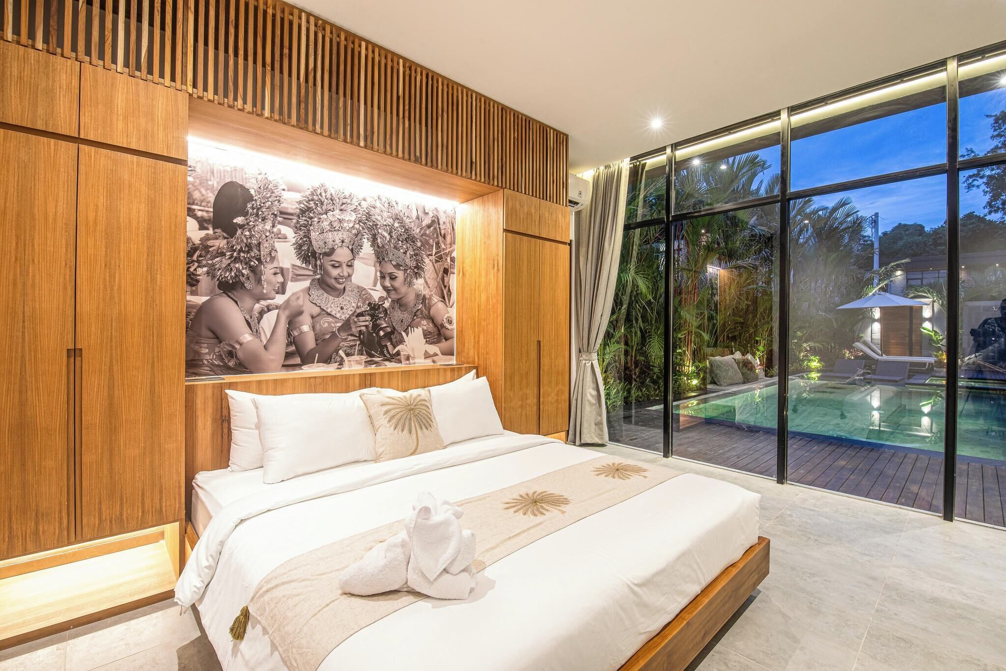 A modern bedroom with a large bed, a wooden panel wall, a large window overlooking a pool, and a tropical-themed photograph.