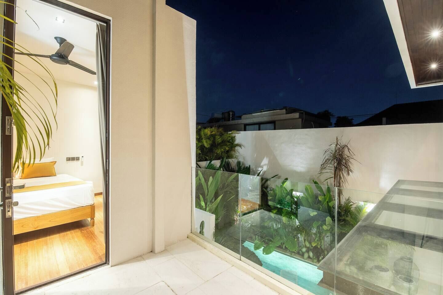 Un balcon moderne doté d’une paroi en verre, un lit avec un matelas blanc et un oreiller jaune, et un ventilateur de plafond.