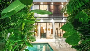 Ett modernt hus med en glasfasad, en pool och frodiga, tropiska växter.