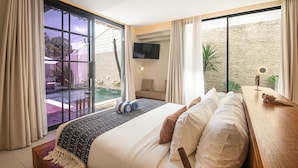 A bedroom with a large bed, a TV mounted on the wall, a small seating area, and a view of an outdoor pool. - Elegant 4BR Seminyak Villa 10min From the Shore (Seminyak)
