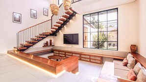 A spacious living area with a wooden staircase, a large window, a sofa, and a TV mounted on the wall. - Elegant 4BR Seminyak Villa 10min From the Shore (Seminyak)