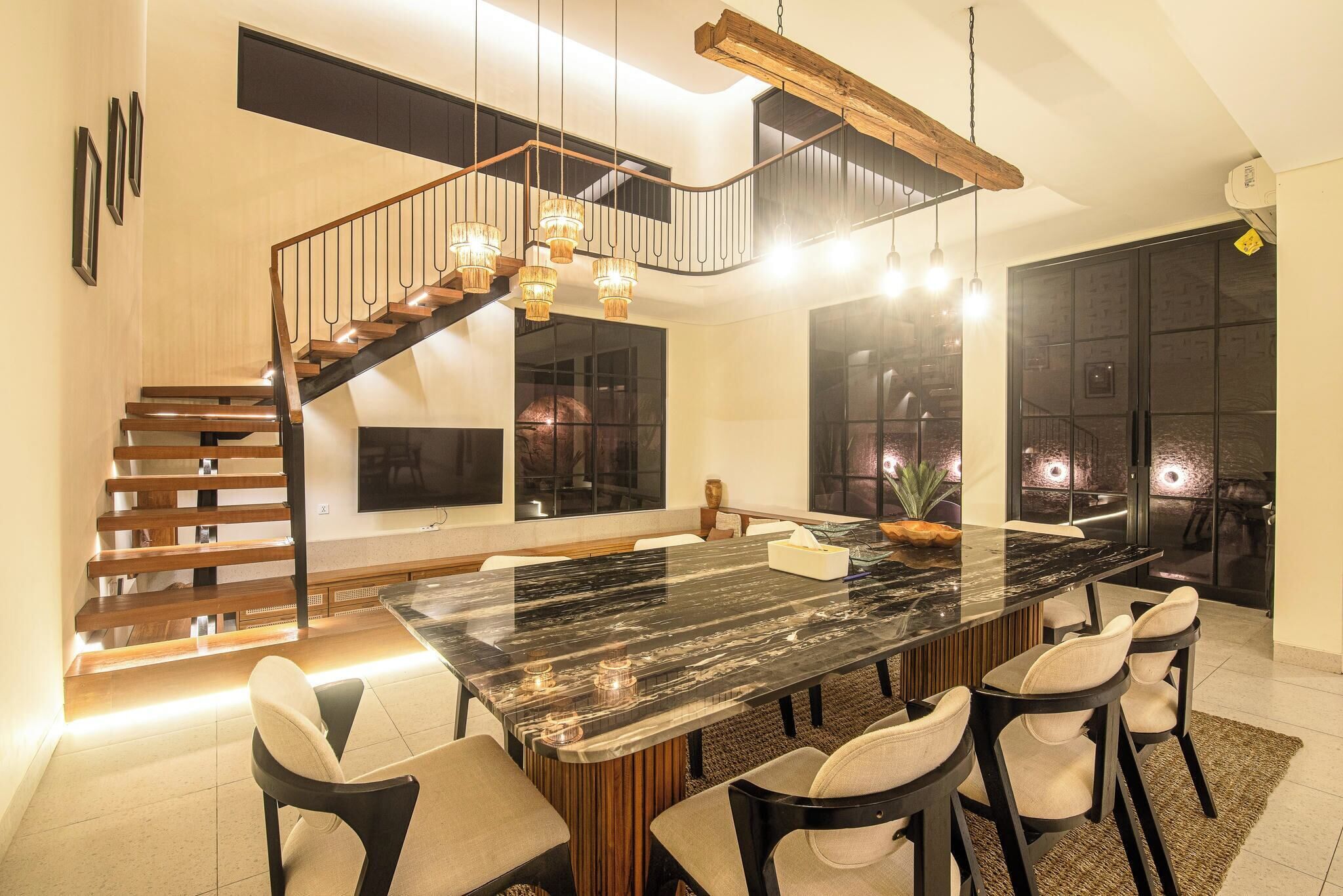 A modern dining area with a large marble table, six chairs, and a staircase with wooden railings.
