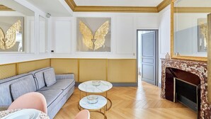 Een kamer met een grijze bank, twee ronde tafels met decoratieve objecten, een open haard en gouden wanddecoraties.