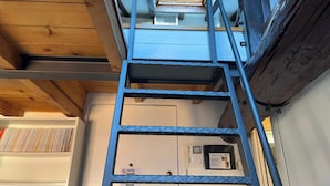 Escadaria de metal que leva a um quarto com parede branca, um telefone e uma pequena janela.