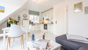 A modern kitchen with white cabinets, a dining area with a white table and chairs, and a gray sofa in a bright, well-lit room. - Syltperle, Germany (Sylt)