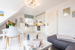 A modern kitchen with white cabinets, a dining area with a white table and chairs, and a gray sofa in a bright, well-lit room. - Syltperle, Germany (Sylt)