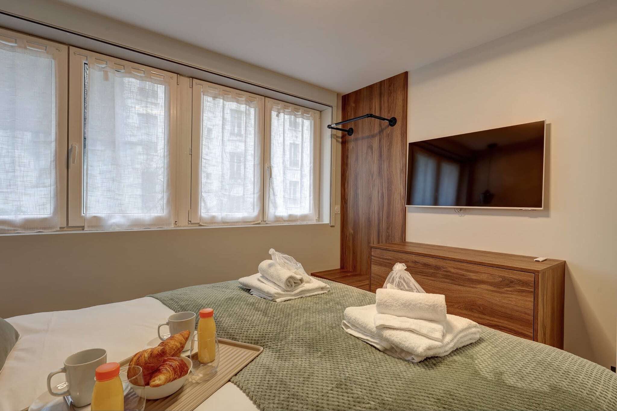 Une chambre d’hôtel avec un lit, une tête de lit en bois, une fenêtre avec des rideaux, un porte-serviettes et un plateau avec des articles pour le petit-déjeuner.
