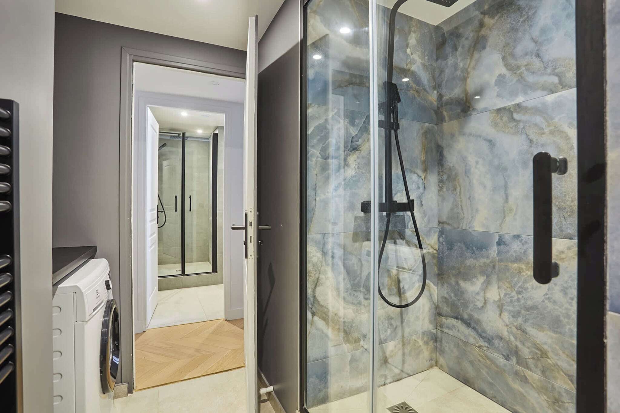 A modern bathroom with a glass shower enclosure, a built-in washing machine, and a glass door leading to another room.