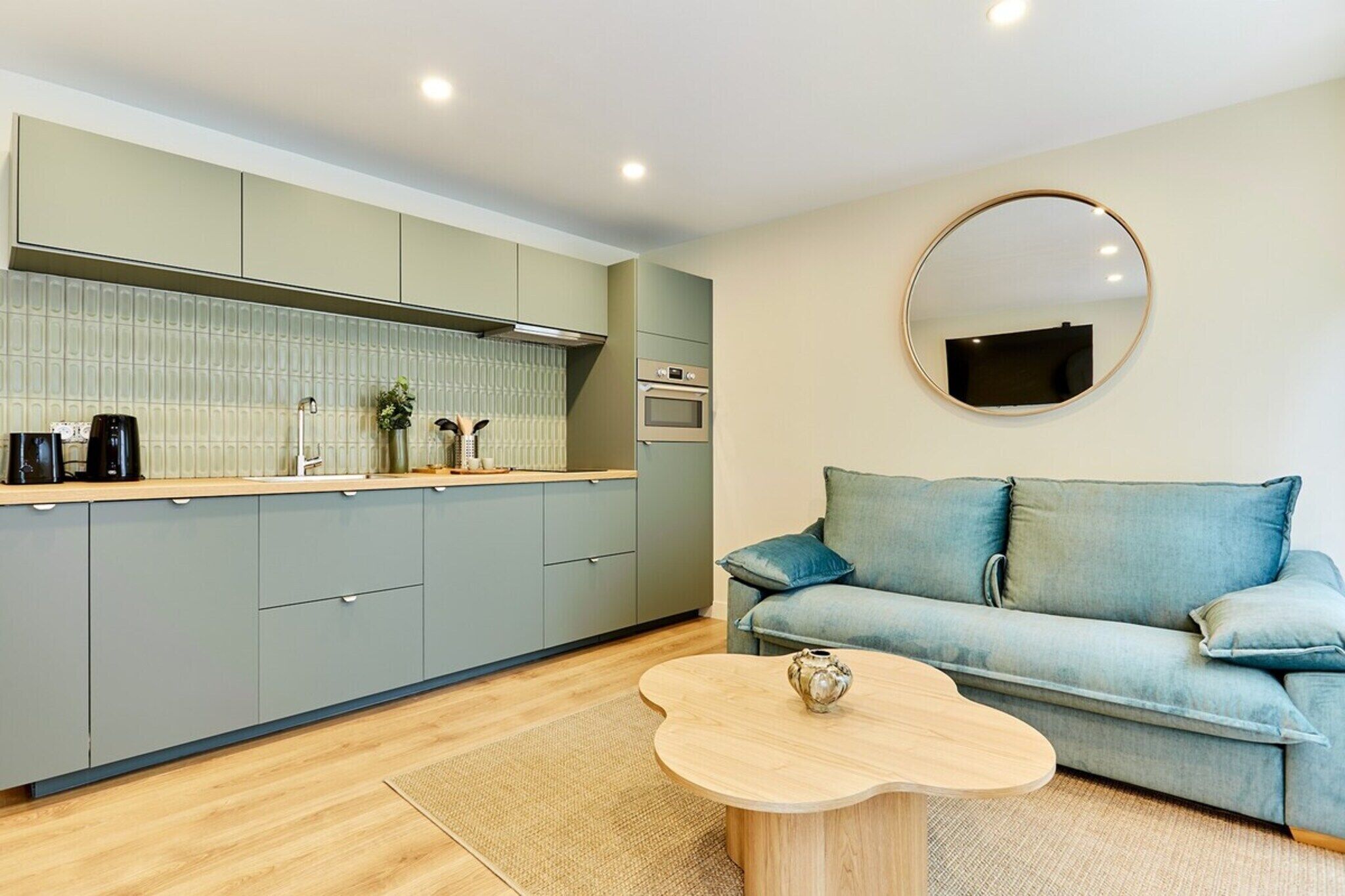 A modern kitchen with grey cabinets, a round mirror, a blue sofa, and a wooden coffee table.