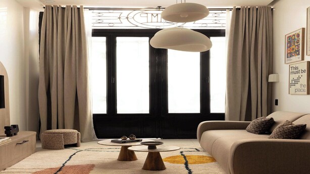 Un salon moderne avec un canapé, une table basse, un pouf et une grande fenêtre agrémentée de rideaux.