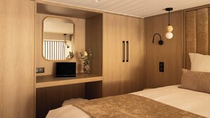 Une chambre d’hôtel moderne avec une armoire en bois, une table de chevet avec un ordinateur portable, un miroir et un lit recouvert d’une couverture à motifs.