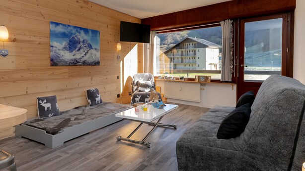 Ett modernt vardagsrum med en soffa, ett litet bord, en stol och ett stort fönster med utsikt över byggnader.