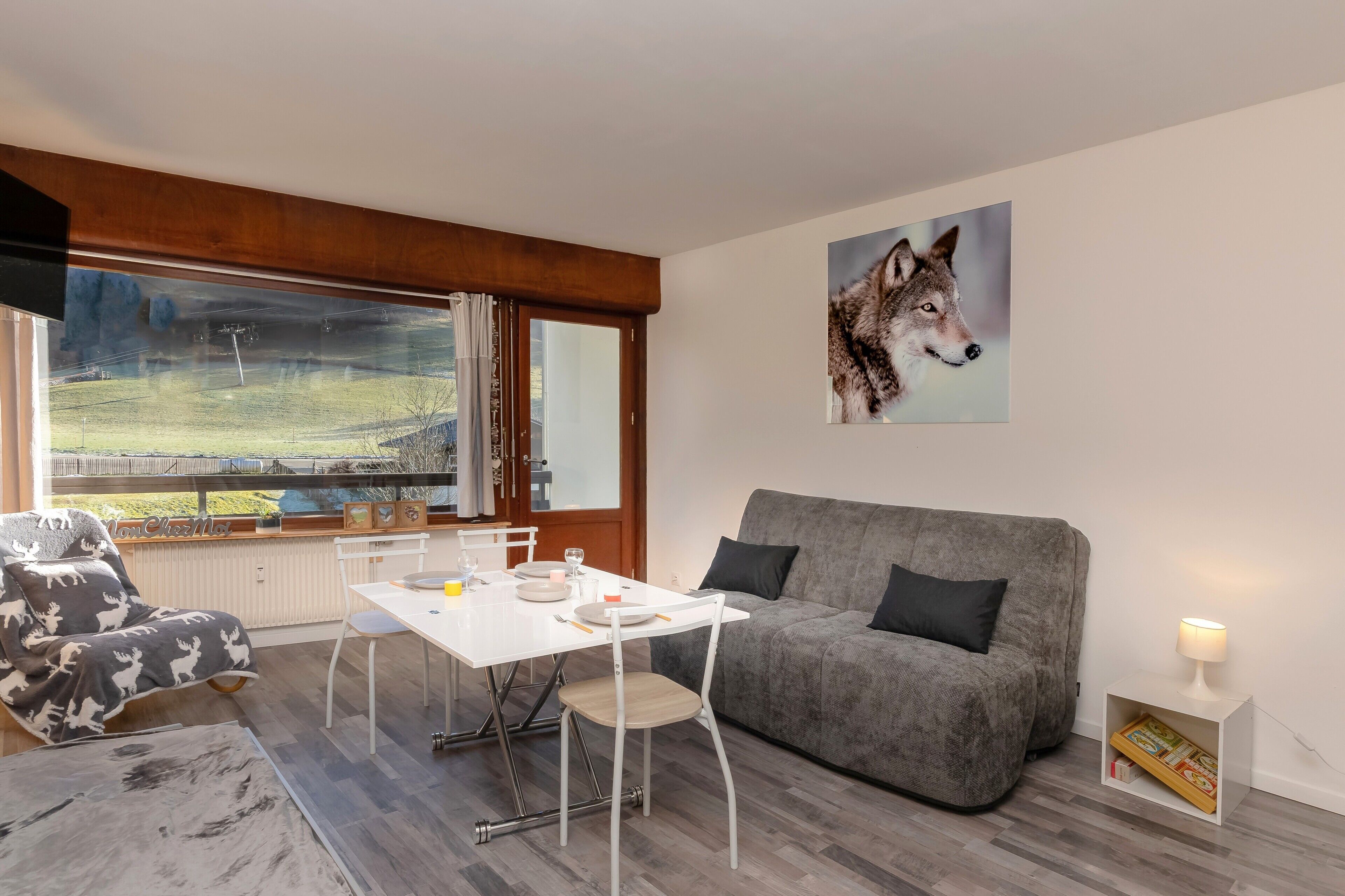 A modern living room with a grey sofa, a white table with chairs, a window with a view of a field, and a wolf painting on the wall.