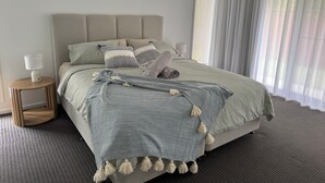 A neatly made bed with a grey and white striped blanket, a grey throw pillow, and a beige throw pillow. There is a small wooden nightstand with a white lamp beside the bed. - The Ponds at Mudgeeraba 4 bedroom Modern House great central location (Mudgeeraba)