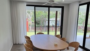 A spacious dining area with a large round table and six chairs, large windows with curtains, and a ceiling fan. - The Ponds at Mudgeeraba 4 bedroom Modern House great central location (Mudgeeraba)